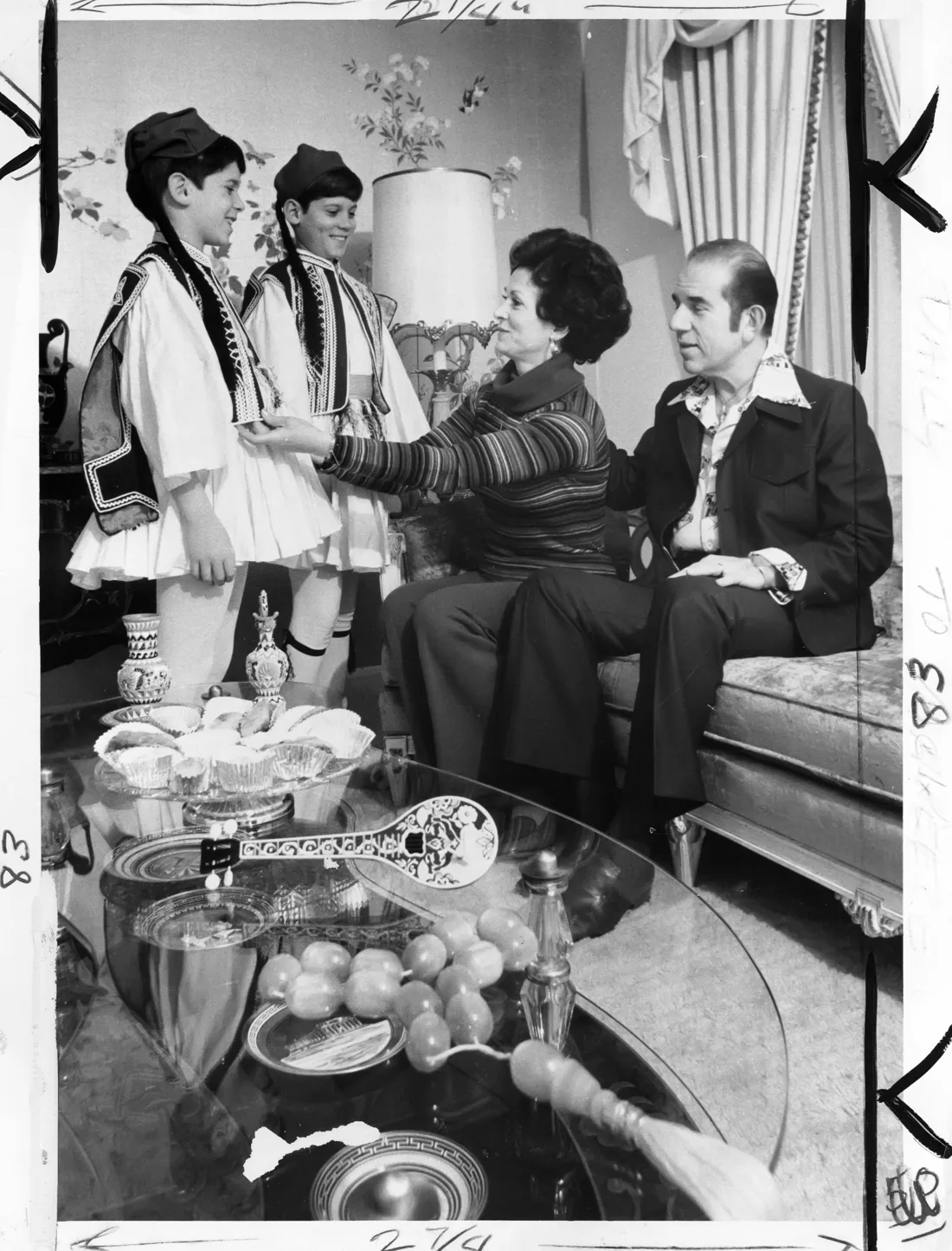 An archival family photograph: parents with two boys in traditional Greek dress, with a bouzouki on the table in front of them