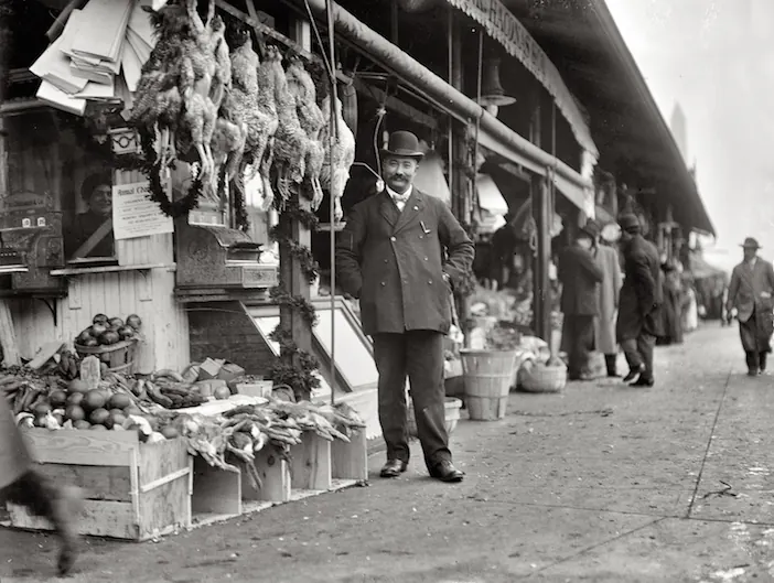 A shopkeeper outside a market with produce and hanging goods